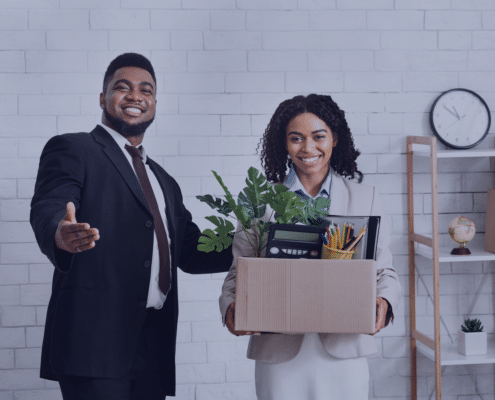 A smiling new employee holding a box of office supplies while being welcomed by a colleague, representing strategies for making a lasting impression in the first 90 days of a new job in 2025.