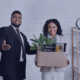 A smiling new employee holding a box of office supplies while being welcomed by a colleague, representing strategies for making a lasting impression in the first 90 days of a new job in 2025.