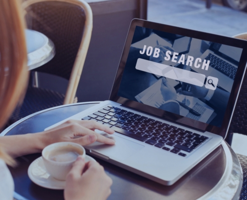 Woman using a laptop with “Job Search” on the screen while holding a coffee cup, symbolizing a mid-year career relaunch through a recruitment agency